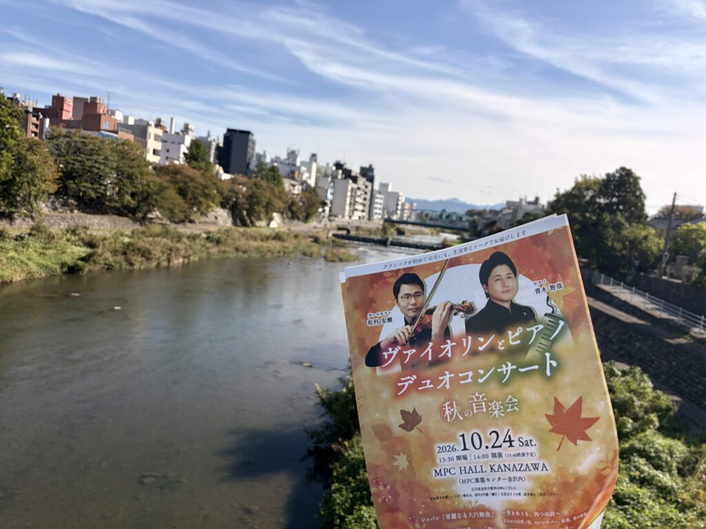 金沢市を流れる犀川を背景に、松村宏樹と青木智哉による2026年10月24日のヴァイオリンとピアノのデュオコンサートのチラシを撮影した写真。